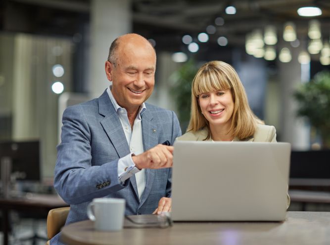 A man and a woman are seated at a table, smiling and looking at a laptop screen. They appear to be in an office setting with blurred background elements.