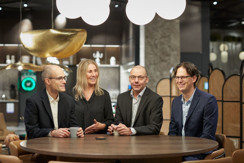 Four people in business attire sit around a wooden table in a modern office setting, smiling and holding coffee cups.